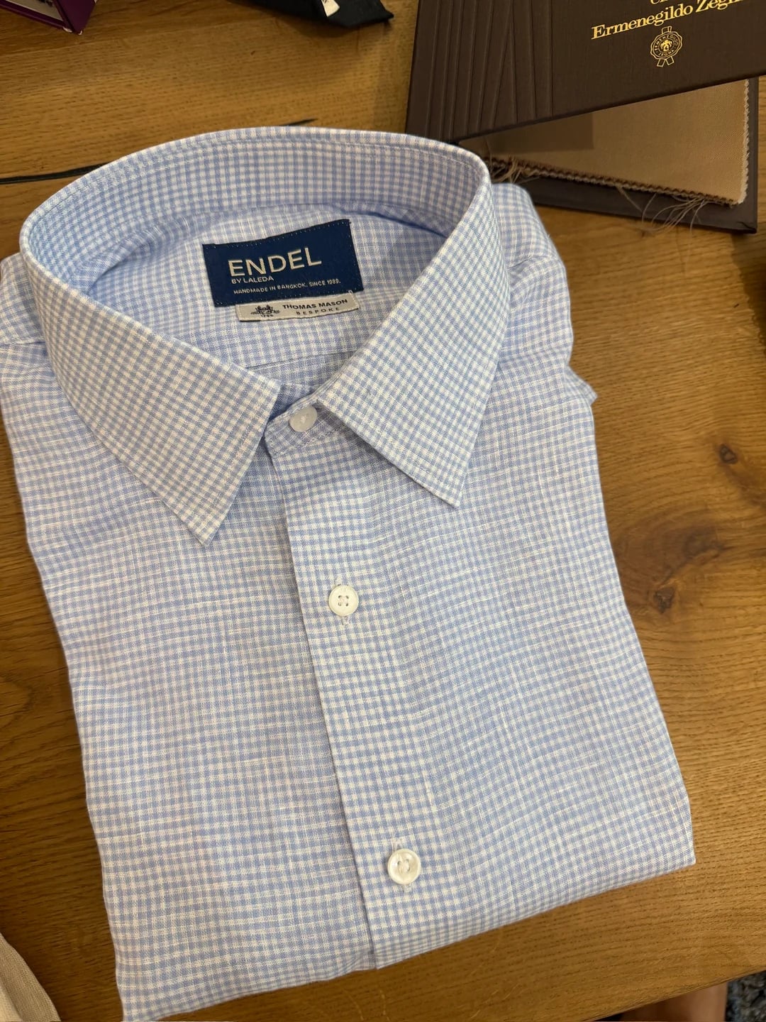 Blaues Gingham-Maßhemd mit Endel-by-Laleda- und Thomas-Mason-Etiketten neben einem Zegna-Stoffbuch bei Endel Tailor Bangkok